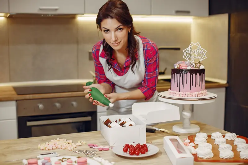 A woman in an apron decorates a cake with icing in a contemporary kitchen setting