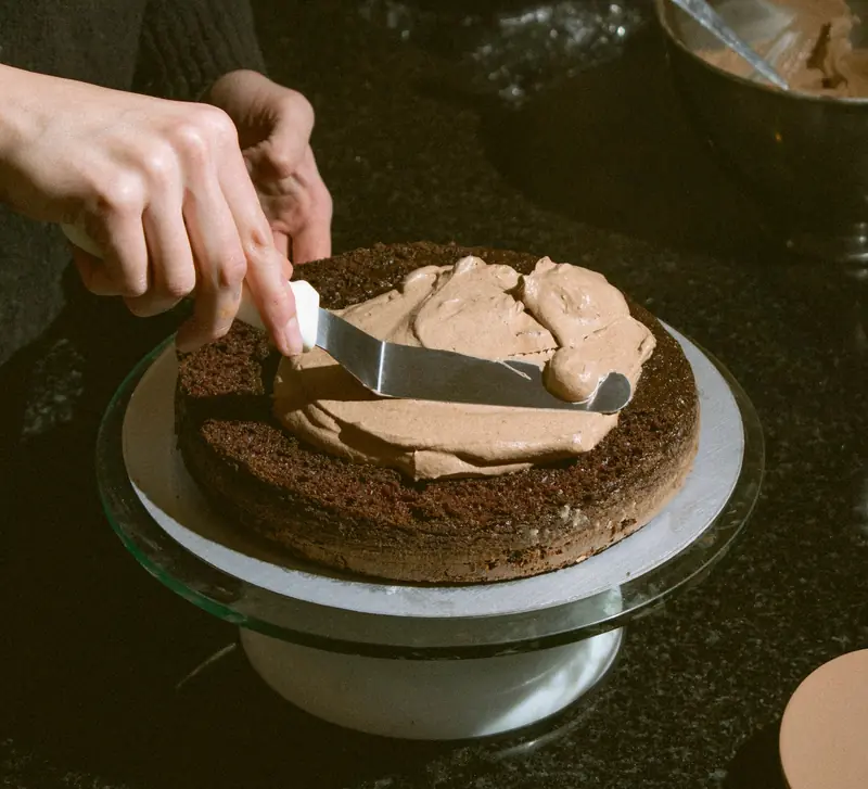 A person decorates a chocolate cake with chocolate frosting on a turntable in a kitchen