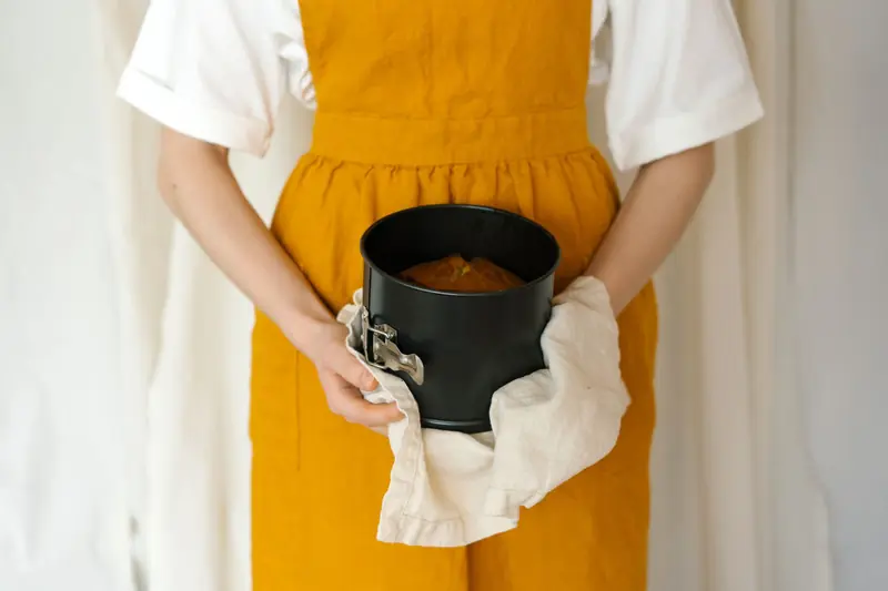A woman in a yellow apron holds a freshly baked cake in a baking pan using a cloth