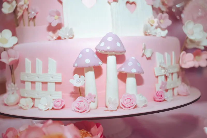 A delightful pink cake with fondant mushrooms and flowers, perfect for whimsical events or fairy tale themes