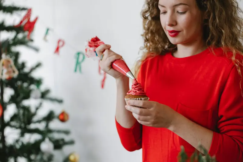 Concentrated young lady in red sweater piping red cream on cupcake with pastry bag in light room with white wall and decorated Christmas tree