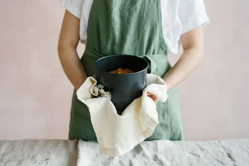 A person in a green apron holds a round baking pan with a cake inside, using a towel