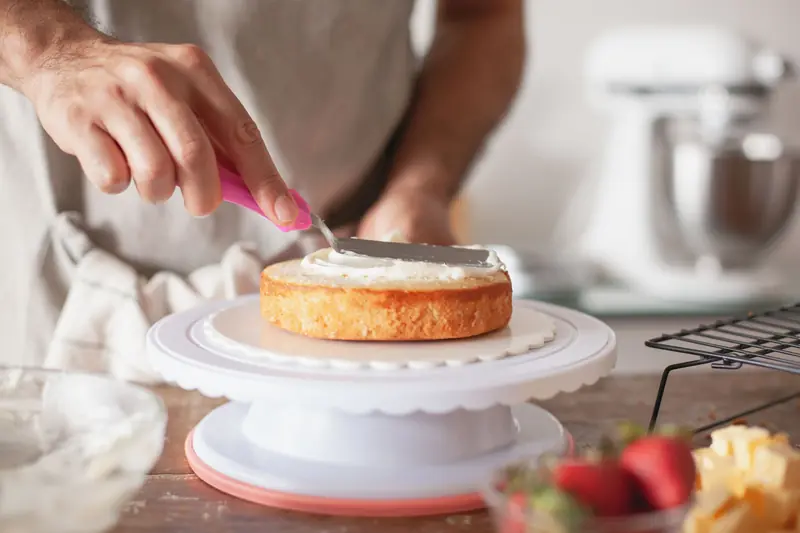 Close-up of person decorating a cake with frosting using a pink spatula on a cake stand
